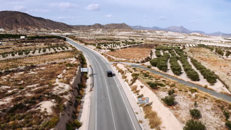 Drone-flying-behind-a-black-SUV,-driving-over-an-asphalt-road-in-the-middle-of-a-dry-grassland-in-Murica,-Spain