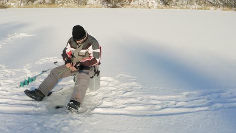 Man-Holding-Rod-Is-Fishing-In-Ice-Hole-On-The-Frozen-Lake-During-Sunny-Wintertime