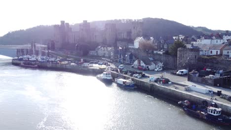 Idyllic-Conwy-castle-and-harbour-fishing-town-boats-on-coastal-waterfront-aerial-lowering-to-water-below