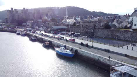 Idyllic-Conwy-castle-and-harbour-fishing-town-boats-on-coastal-waterfront-aerial-pull-away-low-angle
