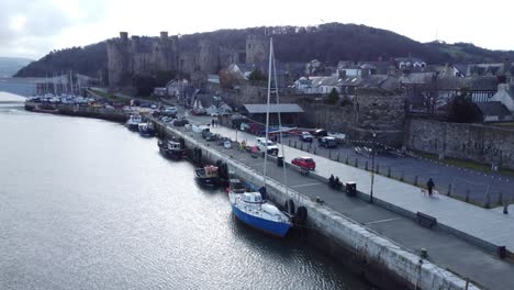 Picturesque-Welsh-Conwy-castle-and-harbour-fishing-town-boats-on-coastal-waterfront-aerial-forward-left-low-angle