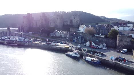 Idyllic-Conwy-castle-and-harbour-fishing-town-boats-on-coastal-shimmering-waterfront-aerial-slow-push-in