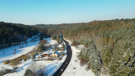 Aerial-wide-shot-showing-cars-on-road-surrounded-by-forest-winter-landscape-with-snow-and-small-house-on-roadside