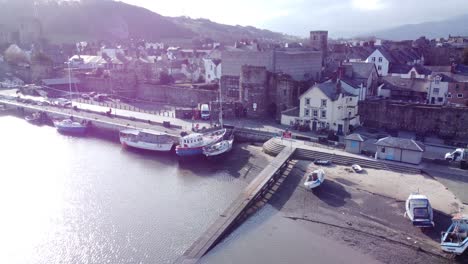 Idyllic-Conwy-castle-and-harbour-fishing-town-boats-on-coastal-waterfront-aerial-left-dolly-over-pier