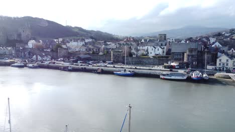 Idyllic-Conwy-castle-and-harbour-fishing-townscape-on-coastal-waterfront-aerial-reversing-over-boats-below
