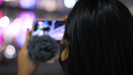 Slow-motion-close-up-head-shot-with-bokeh-of-a-young,-Asian-female-tourist-wearing-a-face-mask,-vlogging-with-her-smartphone-and-speaking-into-a-mic-in-a-city-at-night