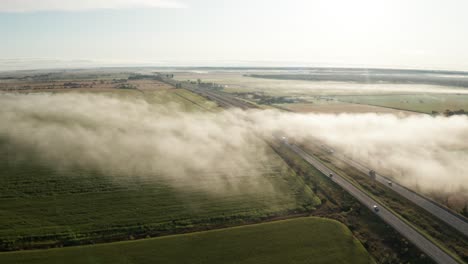 Low-clouds-and-fog-in-early-morning-sunlight-hang-over-fields-and-a-road-with-commuter-traffic-seen-from-drone-flying-overhead