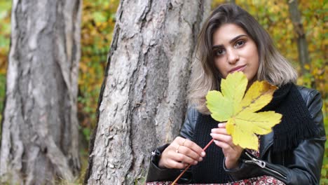 Brunette-girl-in-the-autumn-forest-holds-a-yellow-leaf-in-her-hand-and-spins-it-near-her-face
