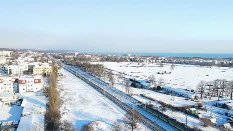 Long-passenger-train-driving-on-a-railway-track-in-a-winter-landscape-with-buildings-covered-with-white-snow-on-a-sunny-day-in-Gdansk-Poland
