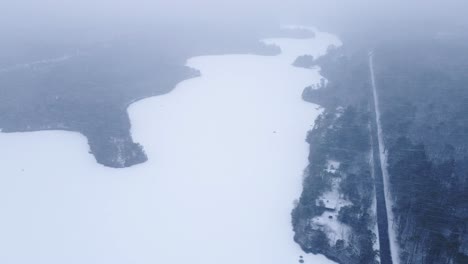 Heavy-Snowstorm-Above-Frozen-Lake-With-Aerial-View-Of-Pine-Trees-And-Asphalt-Road-In-Winter