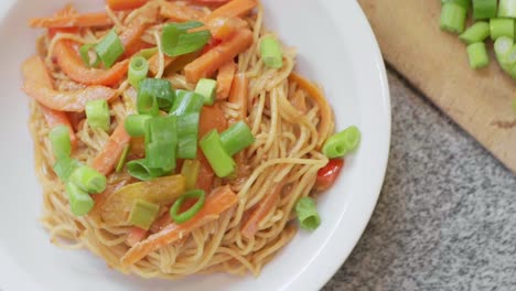 Spaghetti-noodles-with-colorful-sliced-cooked-peppers,-carrots-and-fresh-leek-with-peanut-sauce-on-a-white-plate-steaming-with-more-sliced-leek-on-a-wooden-board