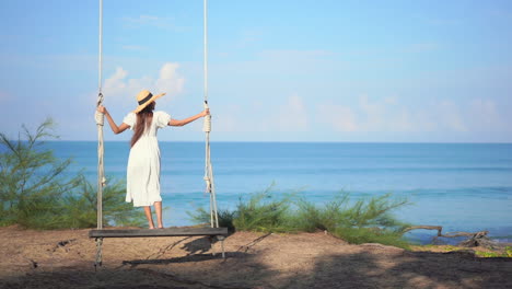 Back-to-the-camera,-A-young-woman-in-a-sundress-and-straw-hat-stands-on-a-large-wooden-swing-overlooking-the-ocean-horizon