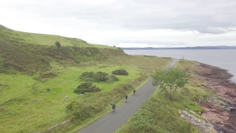 Three-people-cycling-on-a-bike-over-a-black-asphalt-roat-between-the-green-meadows-and-lovely-view-at-Great-Cumbrae,-Scotland-on-a-cloudy-day