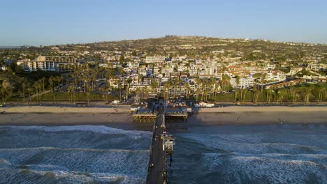 Aerial-flyover-San-Clemente-Pier-with-jetty,-sandy-beach-and-beautiful-cityscape-on-hill