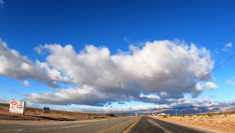 Conduciendo-A-Través-Del-Desierto-De-Mojave-Con-Grandes-Formaciones-De-Nubes-En-Lo-Alto-Y-Las-Escarpadas-Montañas-En-La-Distancia---Punto-De-Vista