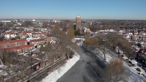Sideways-aerial-urban-winter-scene-with-people-ice-skating-along-the-curved-frozen-canal-going-through-the-Dutch-city-of-Zutphen-with-shadows-of-barren-trees-and-former-water-tower-in-the-background