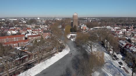 Descending-aerial-snowy-winter-scene-with-people-ice-skating-along-the-curved-frozen-canal-going-through-the-Dutch-city-of-Zutphen-with-shadows-of-barren-trees-and-former-water-tower-in-the-background