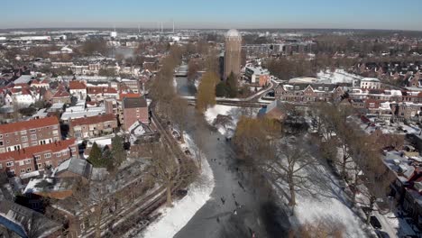 Former-water-tower-aerial-urban-snowy-winter-scene-with-people-ice-skating-along-the-curved-frozen-canal-going-through-the-Dutch-city-of-Zutphen-with-shadows-of-barren-trees-casted-on-the-ice