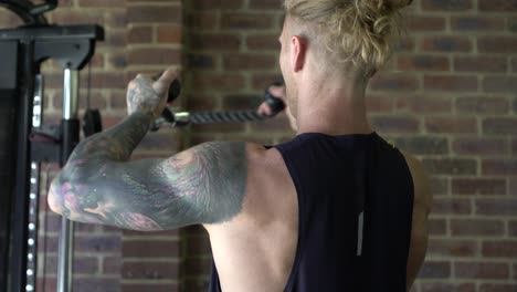 Tattoo-man-muscles-in-home-gym-doing-cable-tricep-over-head-pulls-shot-from-back