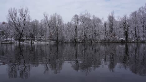 Winter-scene-with-trees-covered-in-snow-reflecting-on-calm-cold-water-of-lake