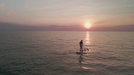 Drone-footage-of-a-young-man-paddle-boarding-in-middle-of-the-ocean-during-sunset,-golden-sunset-in-the-evening-from-the-ocean
