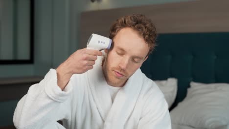 A-young-man-in-a-bathrobe-will-measure-his-temperature-with-a-non-contact-thermometer
