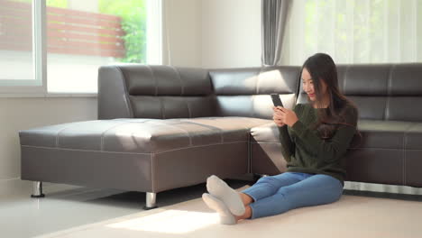 A-pretty-young-woman-sitting-on-the-floor-holding-her-smartphone-laughs-at-what-she-sees