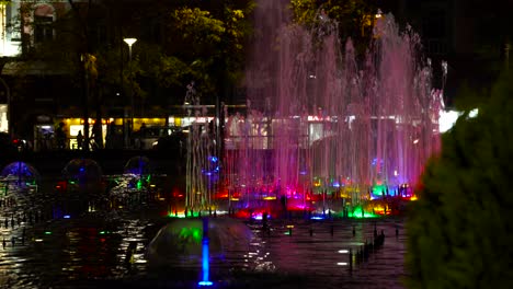 Bunter-Wasserbrunnen-In-Der-Nacht-Mit-Menschen-Und-Autos,-Die-Sich-Auf-Dem-Hintergrund-In-Tirana-Bewegen