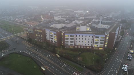 Misty-foggy-hospital-building-UK-town-traffic-aerial-view-rising-tilt-down