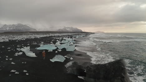 Hombre-Caminando-En-La-Playa-De-Diamantes-De-Arena-Negra-Con-Enormes-Icebergs-Arrastrados-A-Tierra-En-El-Sur-De-Islandia