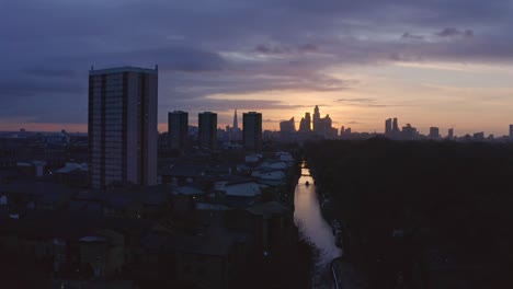 Low-dolly-back-Aerial-drone-shot-of-house-boat-on-London-Canal-Victoria-park-towards-city-skyline-to-highway-at-sunset