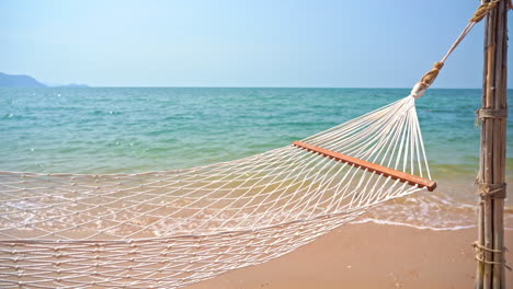 Empty-hammock-on-a-sandy-beach-near-a-calm-ocean-with-waves-slowly-reaching-the-shore-during-hot-sunny-day
