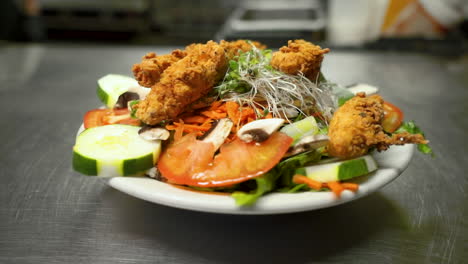 Fried-chicken-strip-salad-sits-on-pass,-vegetables-and-greens-topped-with-chicken,-slide-HD