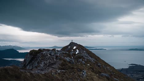 Small-person-standing-with-arms-up-in-the-air-on-top-of-a-large-mountain-peak,-watching-over-a-beautiful-ocean-with-a-grey-sky-in-Norway