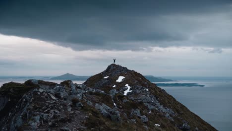 Person-standing-on-top-of-a-large-mountain-with-their-arms-up-in-the-air,-surrounded-by-seascape-in-Norway