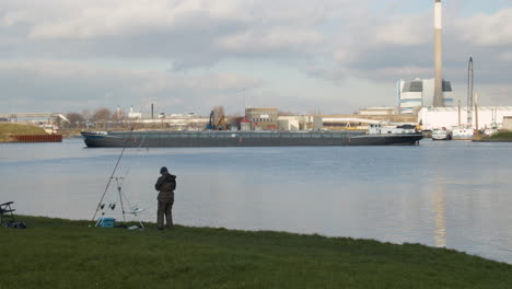 Fisherman-on-shore-preparing-rod-while-a-cargo-ship-moves-over-river