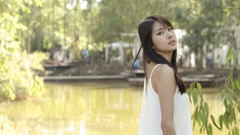 An-Asian-woman-in-a-white-dress-stands-at-the-edge-of-a-pond-and-looks-at-the-camera