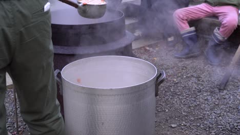 Detail-of-man-pouring-hot-soup-into-big-pot-during-cold-weather-with-people-in-back
