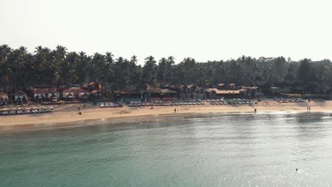 Beach-chairs-resting-in-Palolem-Beach-near-turquoise-ocean-shore---Aerial-Low-angle-Orbit-panoramic-shot
