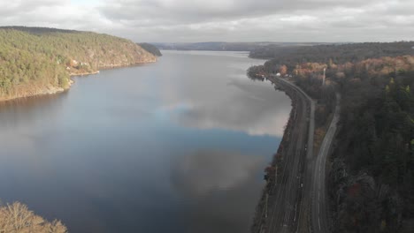 Drone-flying-over-lake-with-cloud-reflections,-road-and-rail-alongside