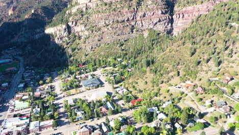 Aerial-Drone-Tilt-Up-of-Beautiful-Ouray-Colorado-Mountain-Town-Surrounded-by-Thick-Pine-Tree-Forest-and-Cliffs-During-Summer-with-Houses-and-Cars-Driving