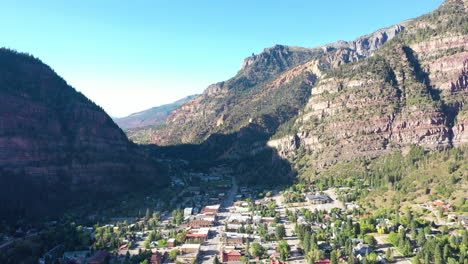 Aerial-Drone-Lowering-Motion-of-Ouray-Colorado-Mountain-City,-Cars-Driving-Through-Downtown-and-Houses-Surrounded-by-Rocky-Mountain-Cliffs-and-Shadow-Cast-Across-Town