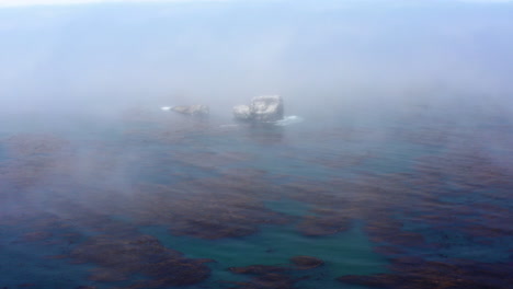 Huge-rocks-sticking-out-of-the-sea,-barely-seen-through-the-mist-that-flows-over-the-water