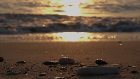 Entspannender-Blick-Auf-Den-Strand-Von-Kieselsteinen-Im-Sand-Mit-Wellen-Im-Hintergrund-Unter-Einem-Goldenen-Sonnenuntergang