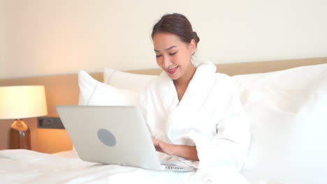 A-pretty-young-woman-works-from-anywhere-on-her-laptop-in-a-comfortable-hotel-bed-surrounded-by-pillows