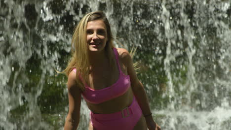 Fun-young-woman-in-a-bikini-in-front-of-waterfall,-smiles-and-splashes-water-at-the-camera,-medium-shot,-slow-motion