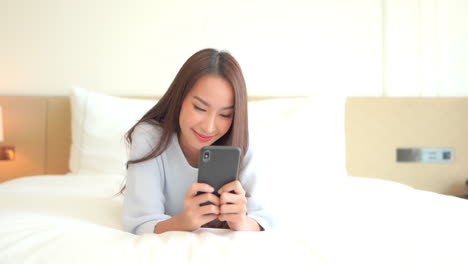 Slow-motion-of-Asian-woman-using-her-phone-with-both-hands-typing-laying-on-the-bed-Hotel-room-wearing-casual-clothes-face-close-up