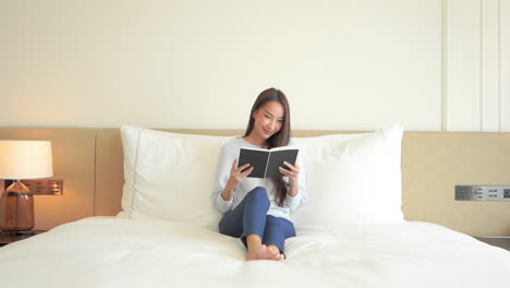 A-young-attractive-woman-enjoys-some-quiet-time-while-sitting-propped-up-in-her-hotel-suite-bed-reading-a-book