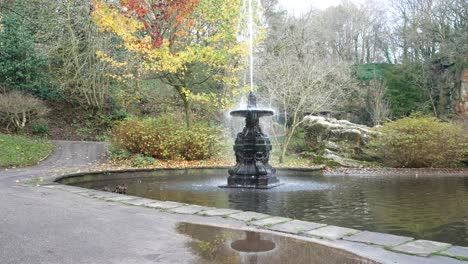 Autumnal-foliage-ornate-English-fountain-sprinkle-spray-in-landmark-historic-garden-landscape