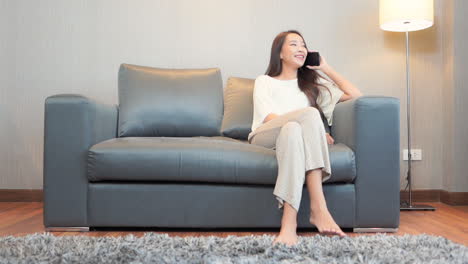 Sexy-Asian-woman-talking-on-a-mobile-phone-while-sitting-on-the-sofa-wearing-casual-clothes-at-home,-flirting-face-expression